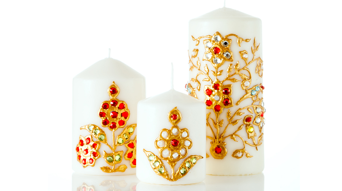 Three white candles of different sizes, each draped with gold flower trimming and decorated with red, silver, and green jewels